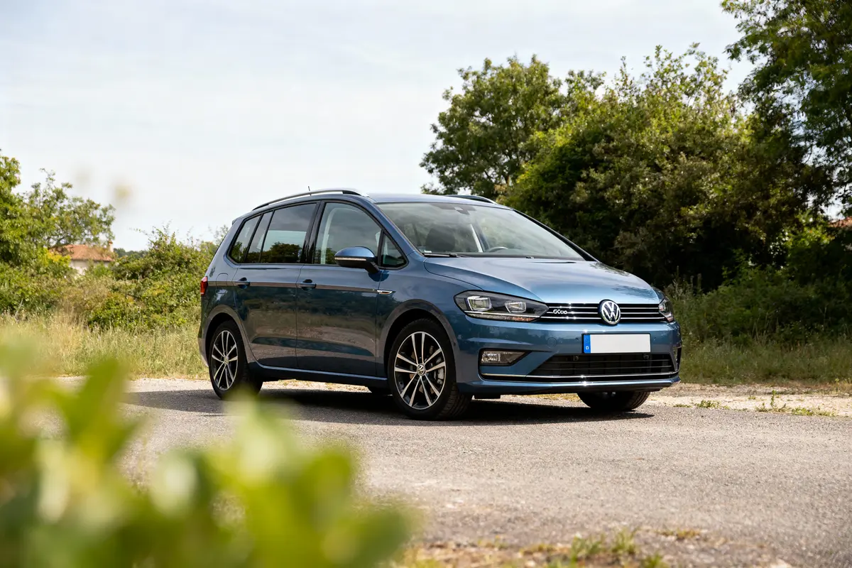 Voiture Volkswagen bleue garée sur une route, entourée de verdure, sous un ciel dégagé.