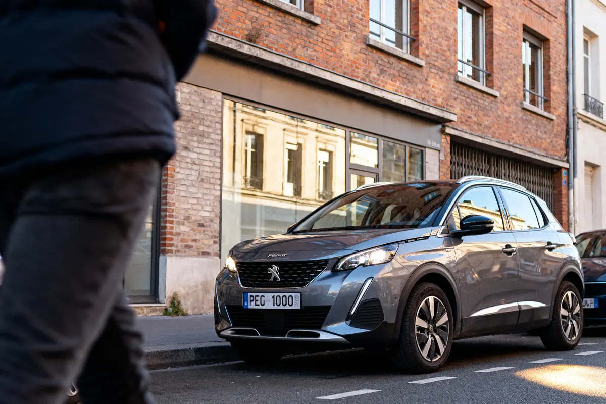 pexels-michal-pech-213601-757318 (1) Voiture Peugeot grise garée devant un bâtiment en briques, avec piéton flou au premier plan.
