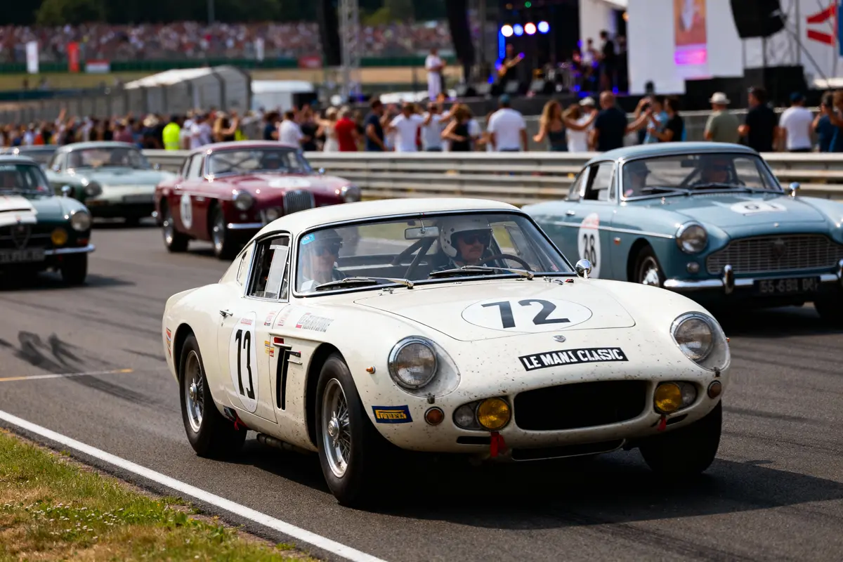 Voitures classiques en course, "Le Mans Classic", participants et spectateurs en arrière-plan, piste de course ensoleillée.