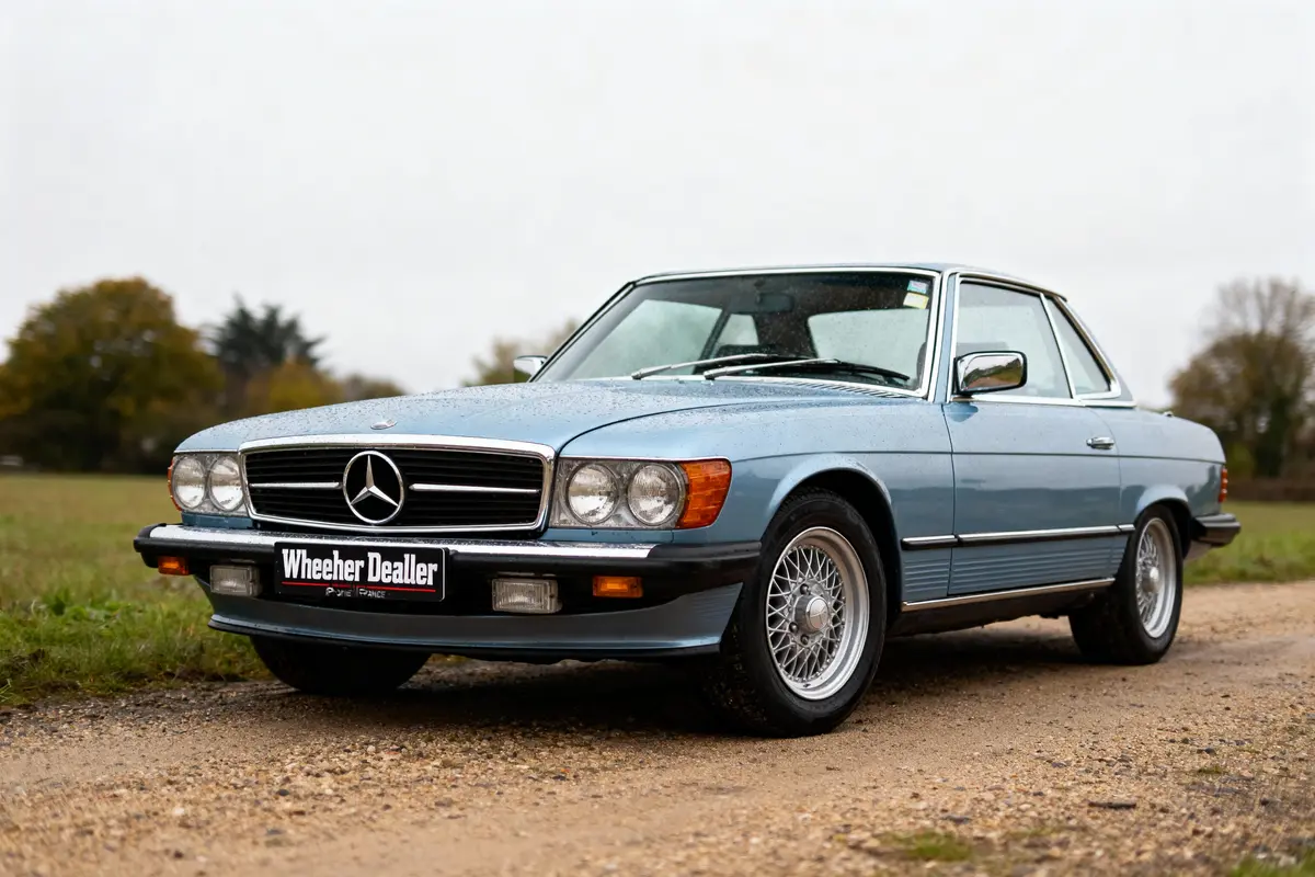 Voiture classique Mercedes bleue stationnée sur un chemin, pare-brise humide, environnement rural. Plaque marquée "Wheeler Dealer".