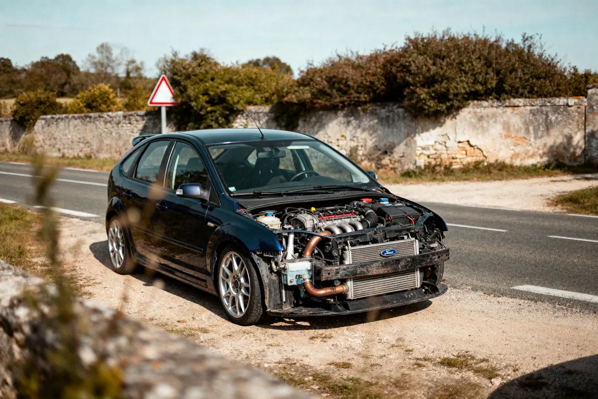 pexels-michal-pech-213601-757318 (1) Voiture noire sans pare-choc avant stationnée sur le bord de la route, devant un mur de pierre.