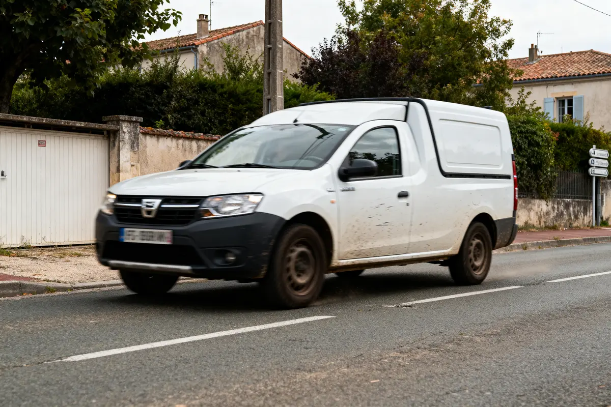 pexels-michal-pech-213601-757318 (1) Utilitaire blanc Dacia roulant sur route, environnement résidentiel verdoyant en arrière-plan. Vitesse modérée.