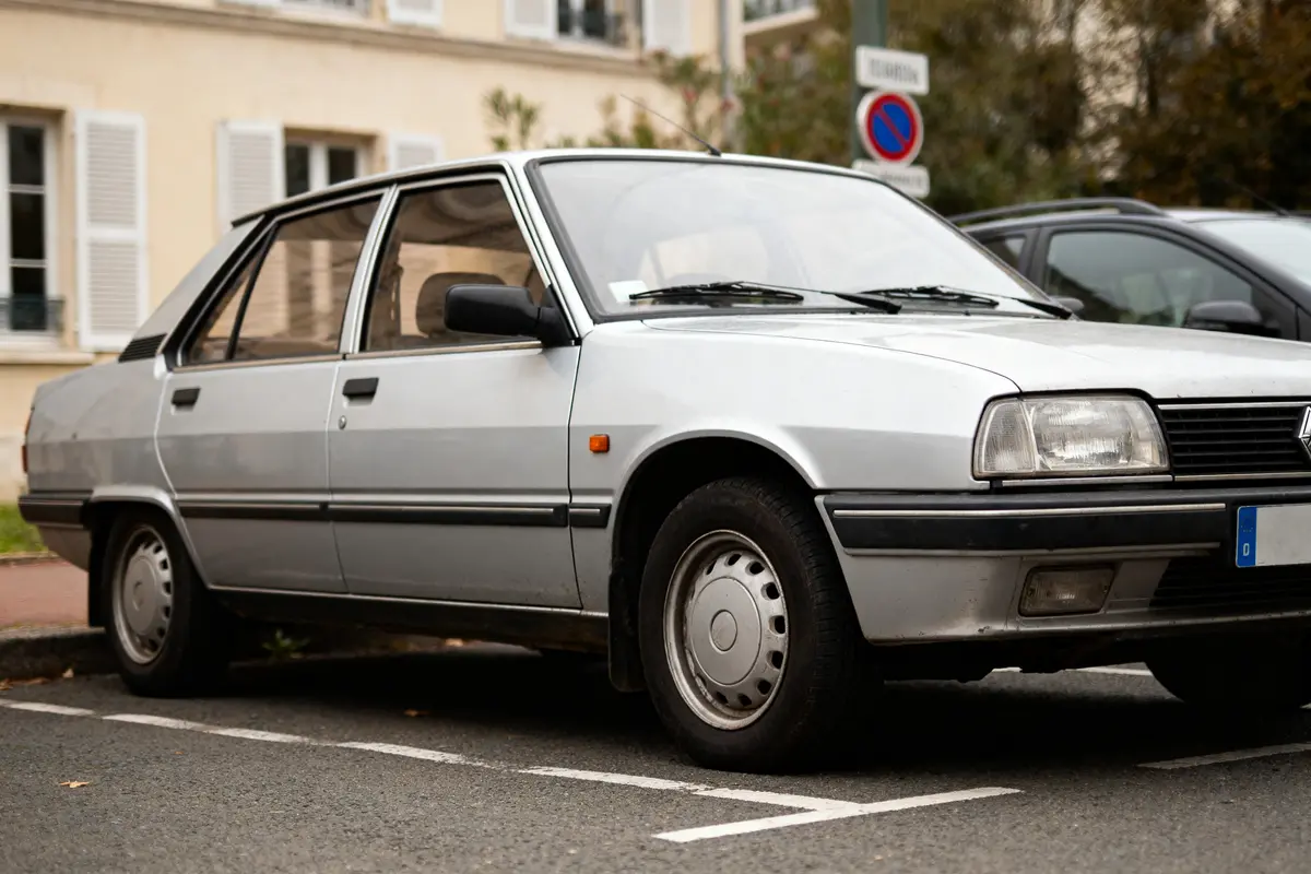 Voiture classique grise, garée silencieusement devant un bâtiment avec fenêtres et volets blancs en arrière-plan.