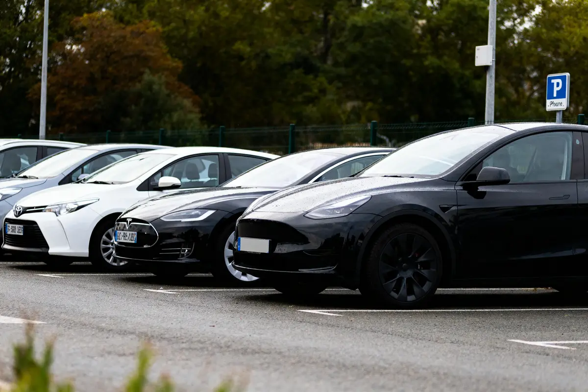 Voitures garées sur un parking, incluant des modèles Tesla et Toyota, sous un ciel nuageux.