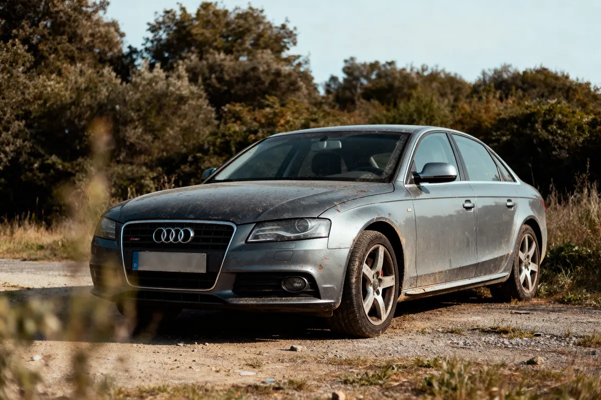 pexels-michal-pech-213601-757318 (1) Voiture Audi grise sale garée sur chemin terreux, arbres en arrière-plan par temps clair.
