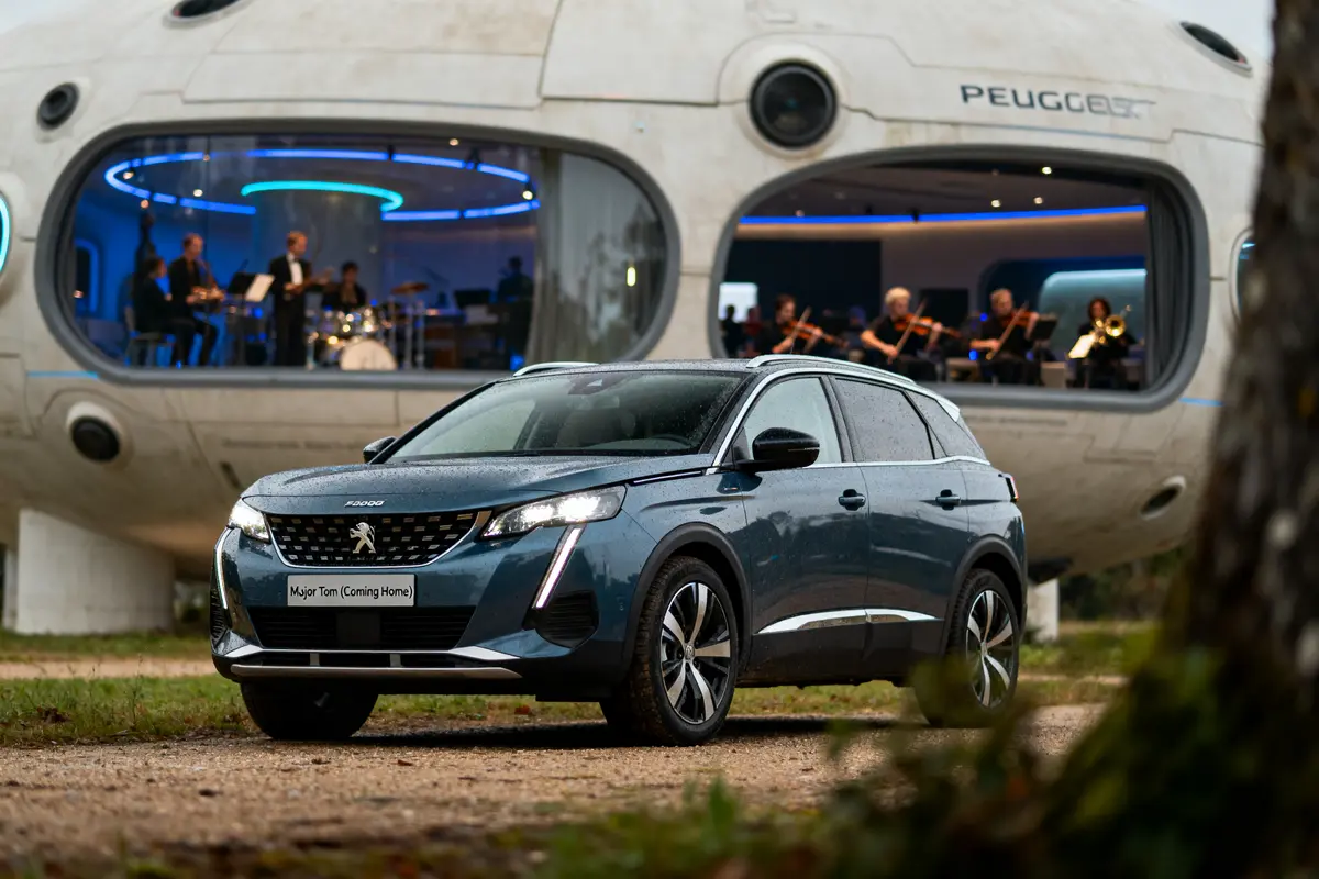 pexels-michal-pech-213601-757318 (1) SUV Peugeot moderne devant un bâtiment futuriste avec musiciens à l'intérieur, éclairage bleu, extérieur pluvieux.
