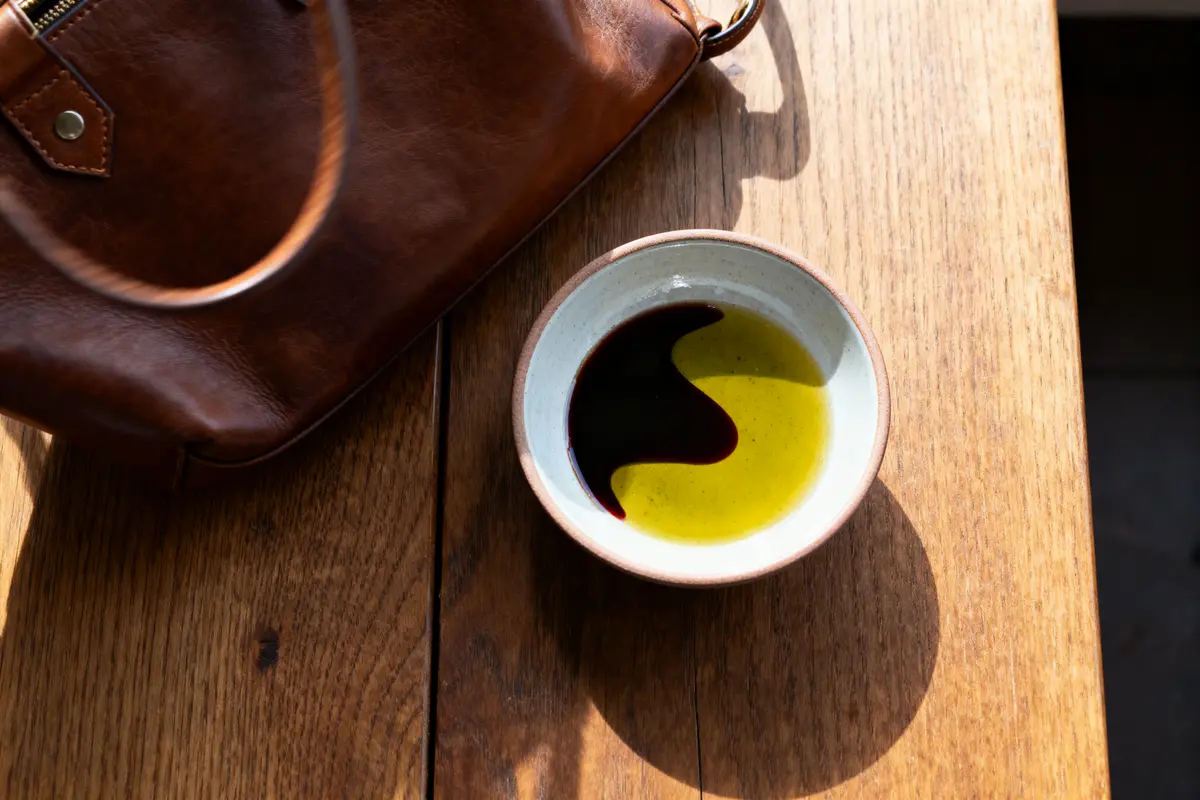 pexels-michal-pech-213601-757318 (1) Bol avec huile et vinaigre sur table en bois, sac en cuir brun à côté, éclairé par le soleil.
