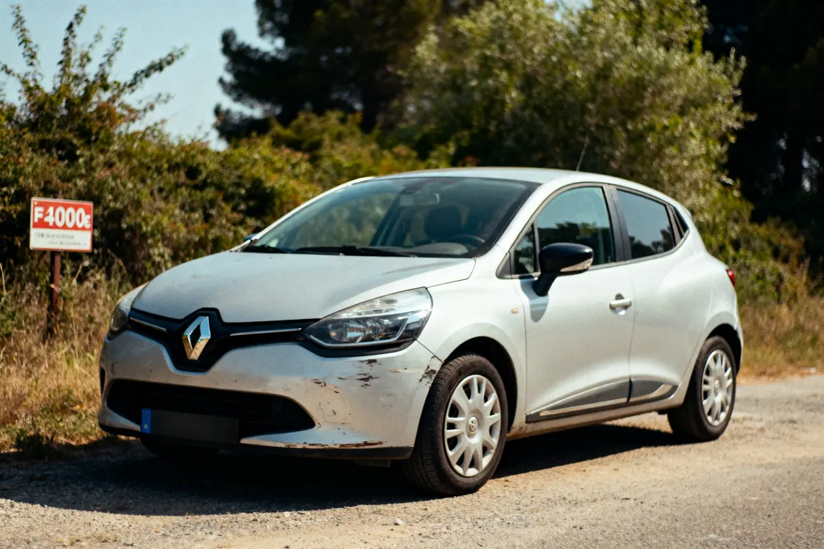 Voiture blanche Renault Clio endommagée stationnée avec panneau indiquant prix de vente 4000 euros. Arrière-plan végétal.