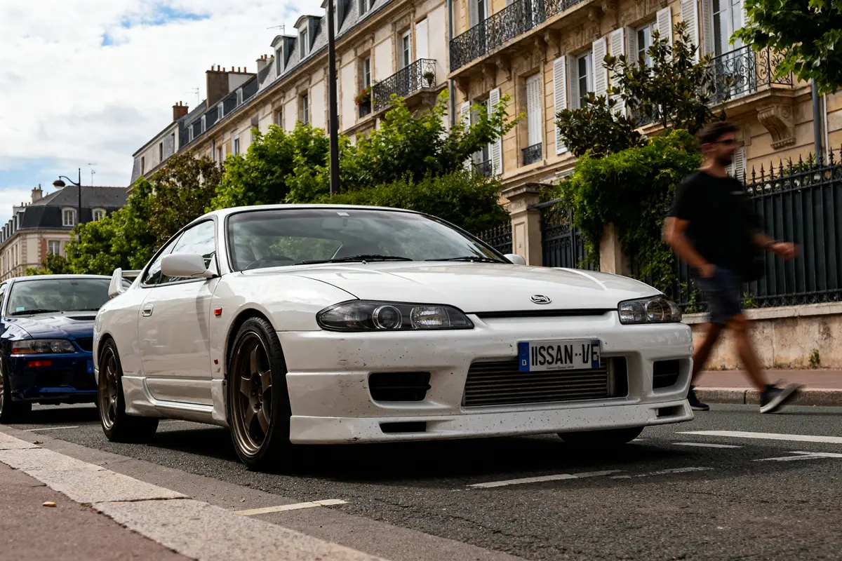 pexels-michal-pech-213601-757318 (1) Voiture blanche Nissan Silvia S15 garée dans une rue résidentielle avec passant flou à côté
