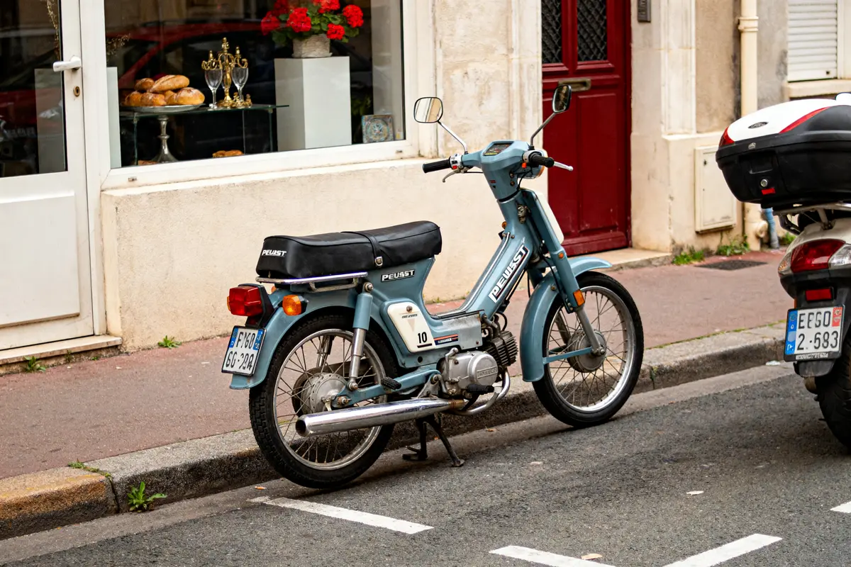 pexels-michal-pech-213601-757318 (1) Scooter Peugeot 105 bleu garé devant une boulangerie avec vitrines remplies de pains.